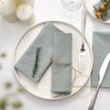 A set of ten light gray cotton table napkins neatly folded on a white plate with silverware and a glass.