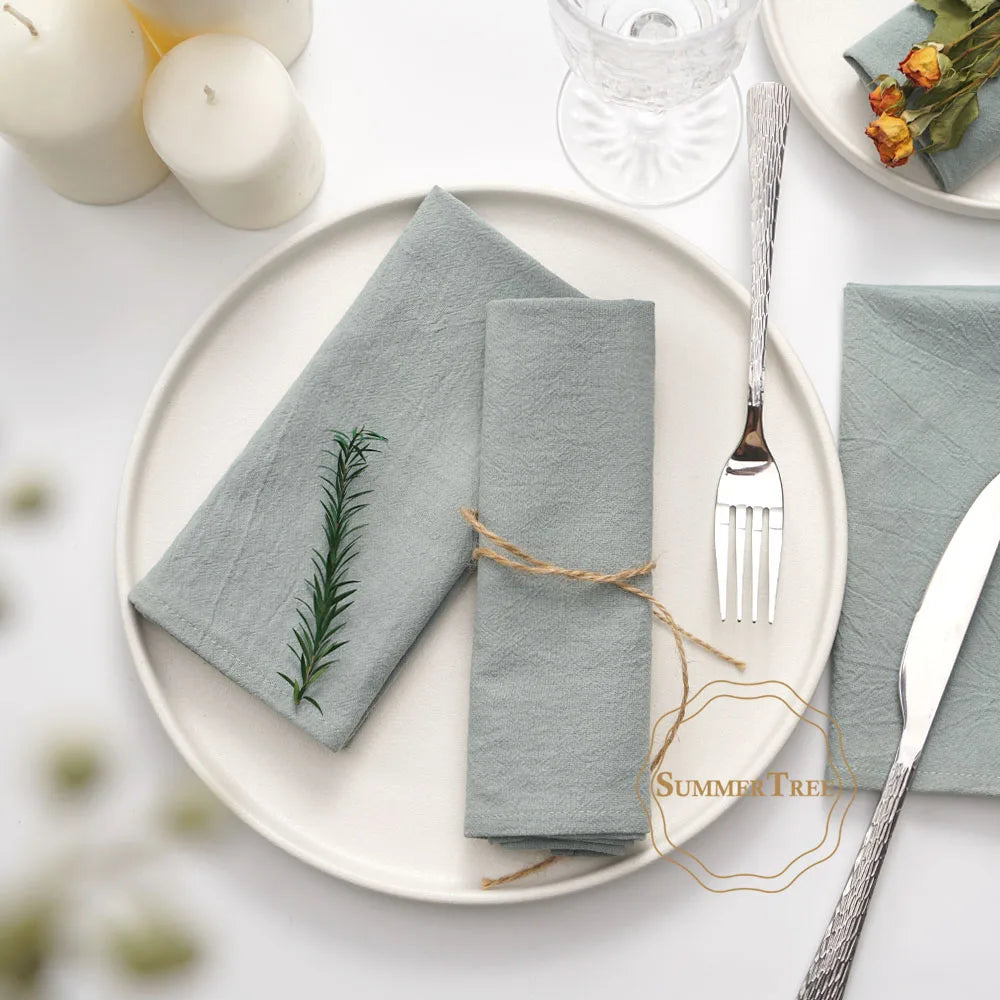 A set of ten light gray cotton table napkins neatly folded on a white plate with silverware and a glass.