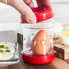 Une main utilise un outil à manivelle rouge pour enlever la coquille d'un œuf dans un récipient transparent.