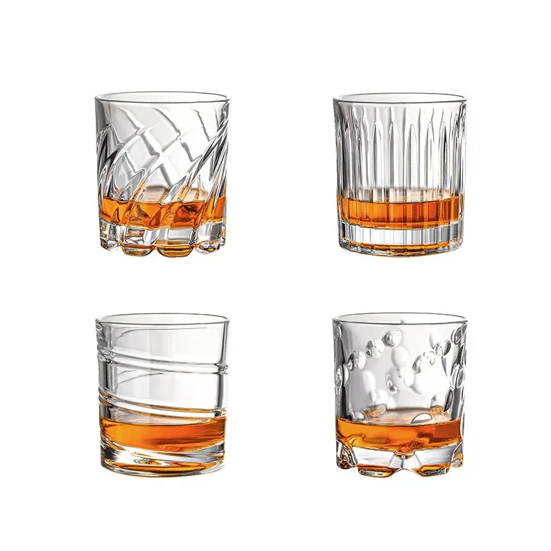 Un juego de cuatro vasos transparentes para whisky, incluyendo uno texturizado y un vaso bajo, llenos de líquido ámbar.
