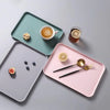 A pink rectangular serving tray holds a glass of juice and two spoons on a gray surface.