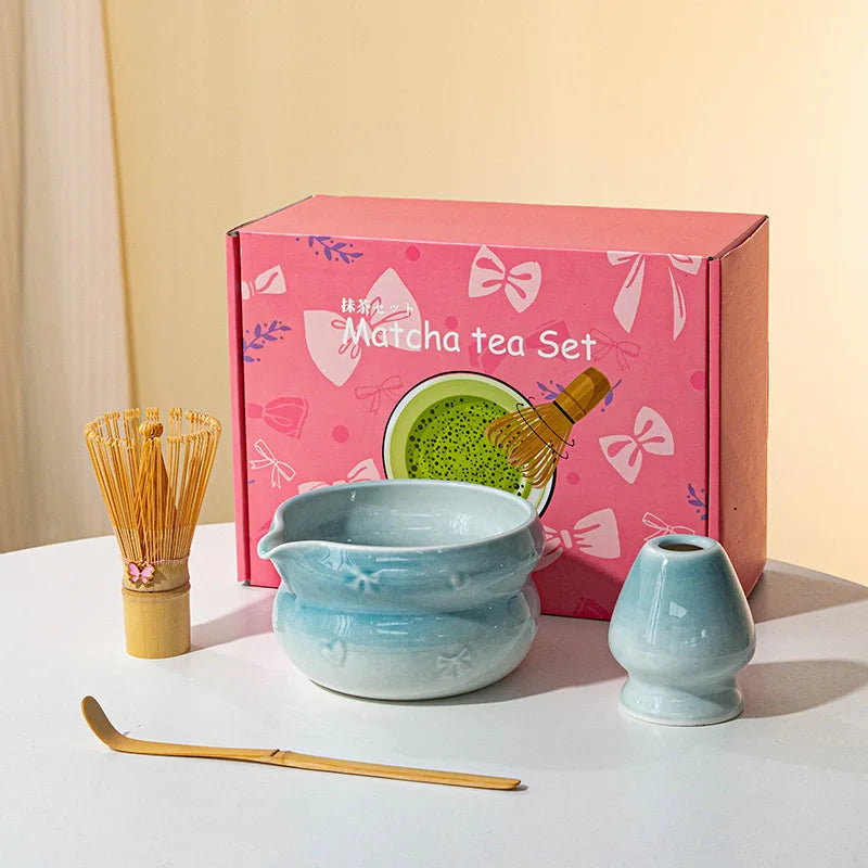 A pink box containing a matcha tea set with a ceramic bowl, bamboo whisk, and spatula on a white surface.