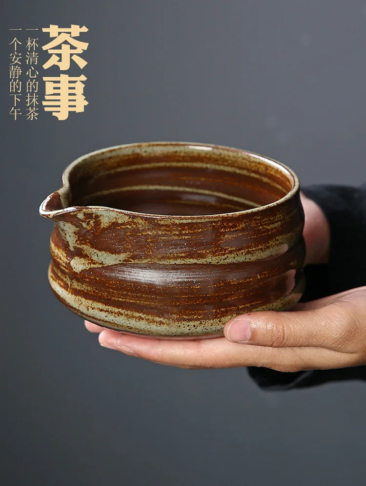 A hand holds a dark brown ceramic bowl, likely part of a Japanese matcha whisk set, against a dark gray background.