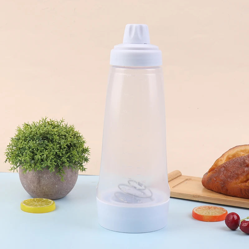 A clear plastic handheld batter mixer bottle with a white cap sits on a light blue surface next to a potted plant and a lemon