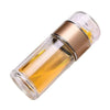 A clear glass tea infuser with a gold-colored cap and a small sprig of dried herb inside.
