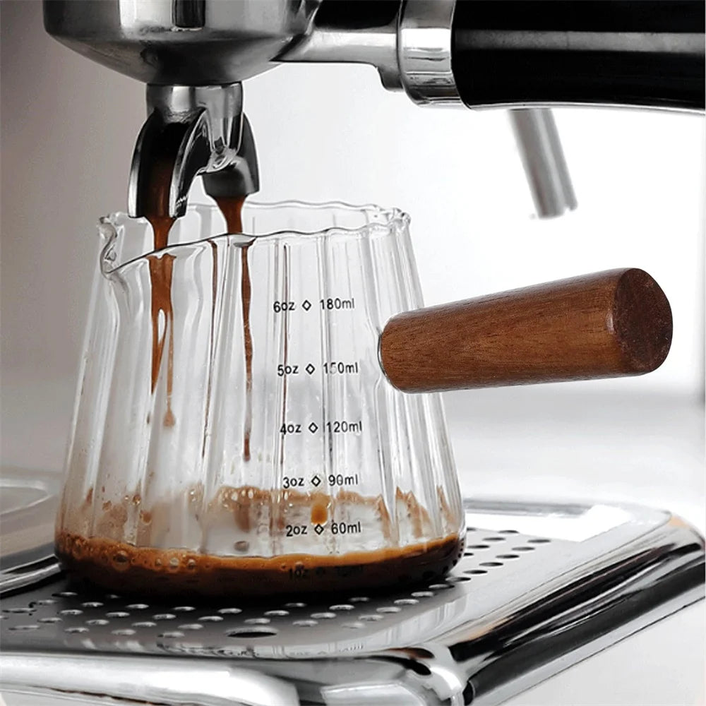 A close-up of espresso shots being poured into glass measuring cups with handles on a metal tray.