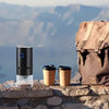 A portable USB-C burr coffee grinder sits on a stone ledge with a cup and a brown backpack in the background overlooking a la
