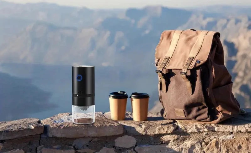 A portable USB-C burr coffee grinder sits on a stone ledge with a cup and a brown backpack in the background overlooking a la