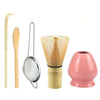 A set of natural bamboo tea tools including a spoon, a strainer, and a bamboo whisk in a pink ceramic cup.