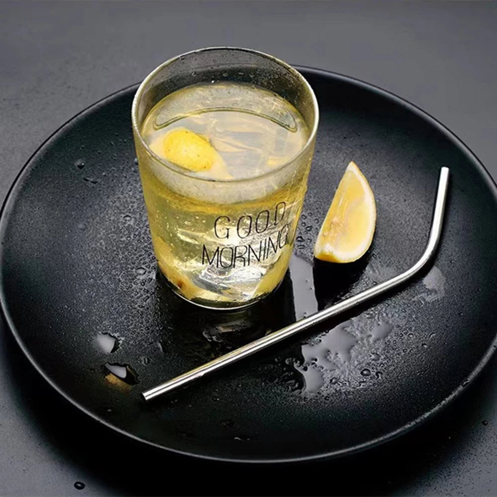 A glass of yellow liquid with lemon slices on a black plate next to a stainless steel drinking straw.