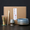 A set of Japanese matcha whisks and a ceramic bowl are displayed in front of a brown cardboard box.