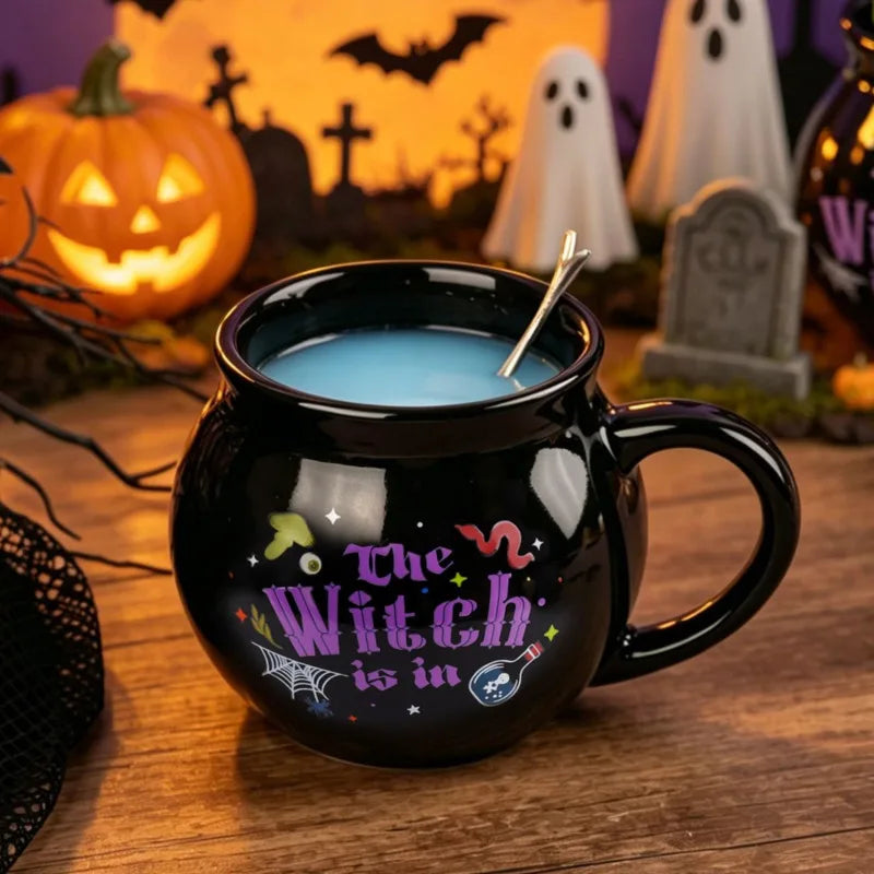 A black cauldron mug filled with blue liquid sits in front of a Halloween-themed background featuring jack-o'-lanterns and gh
