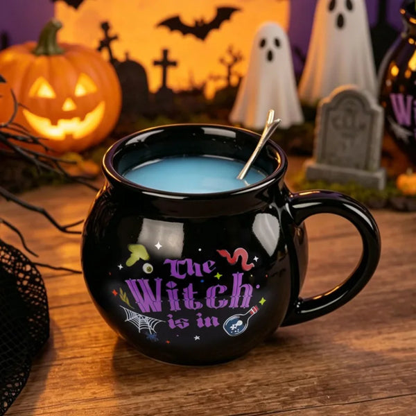 A black cauldron mug filled with blue liquid sits in front of a Halloween-themed background featuring jack-o'-lanterns and gh