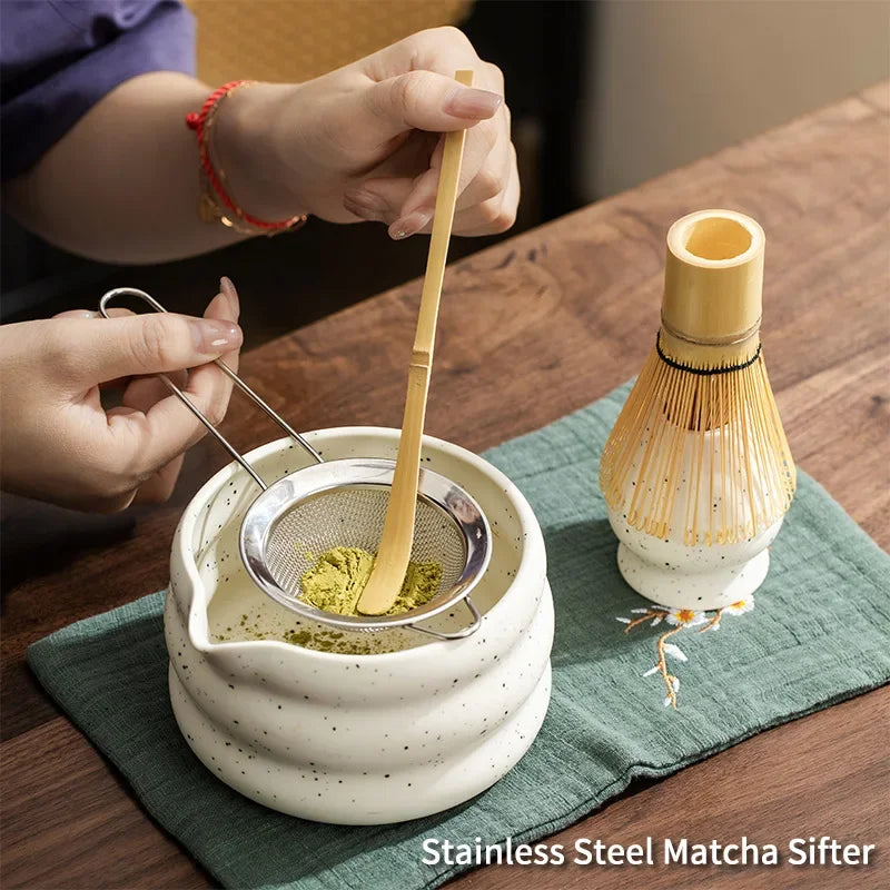 A hand is using a bamboo sifter to scoop matcha powder into a ceramic bowl on a green cloth.