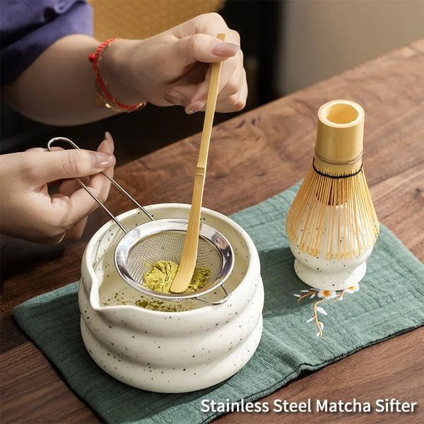 Eine Hand verwendet ein Bambussieb, um Matcha-Pulver in eine Keramikschale auf einem grünen Tuch zu schöpfen.
