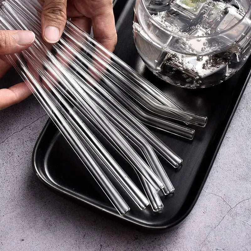 A hand holds a bundle of clear glass drinking straws resting on a black tray next to a glass bowl.