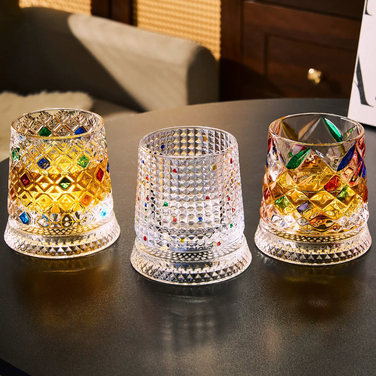 Three rotating whiskey glasses with colorful facets are displayed on a dark surface.