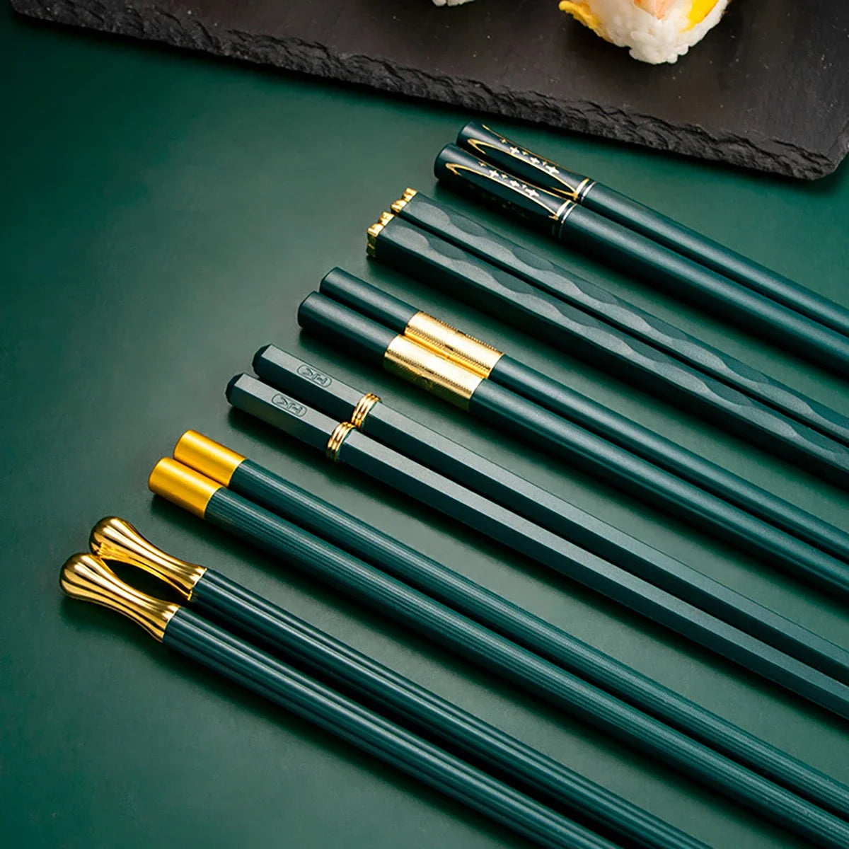 Un juego de palillos reutilizables de aleación verde oscuro y dorado sobre una superficie de pizarra oscura.