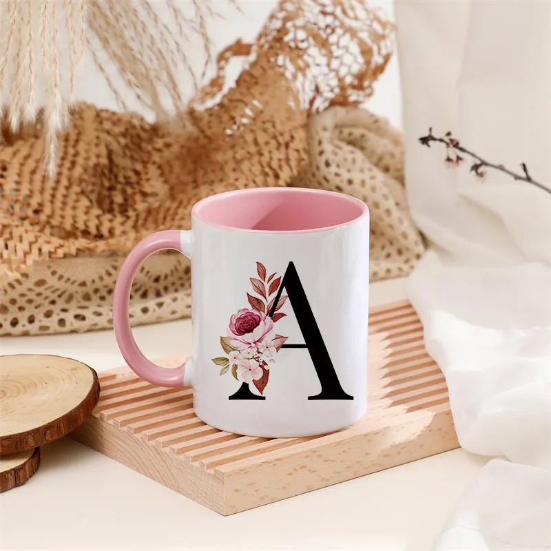 A white coffee mug with a pink handle features a large black letter A monogram in pink and green floral accents.