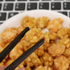A pair of black chopsticks is lifting a golden-brown fried snack from a bowl of similar food.
