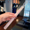 A hand holds a clear plastic chopsticks set with pink tips against a blurred indoor background.