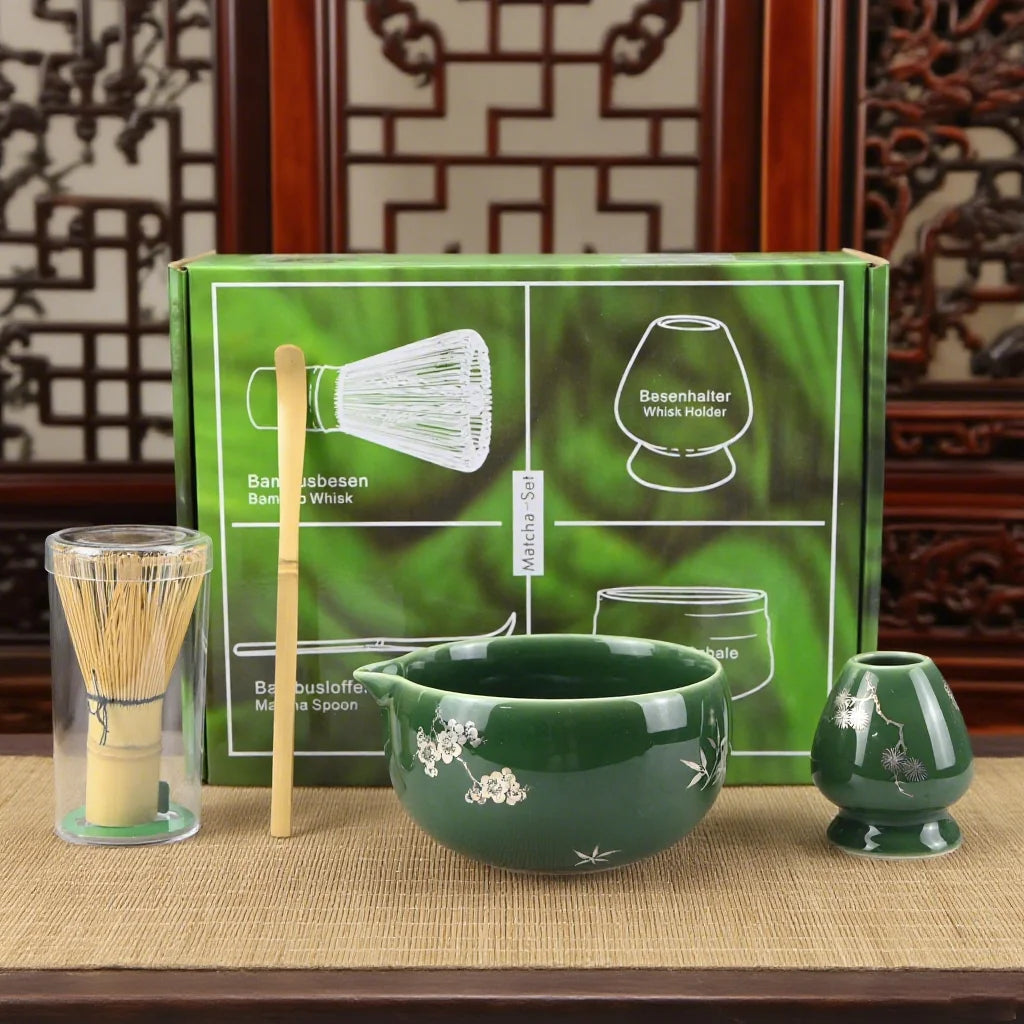 A green box containing a bamboo whisk and a whisk holder, with a bamboo spoon and a green bowl displayed on a wooden surface.