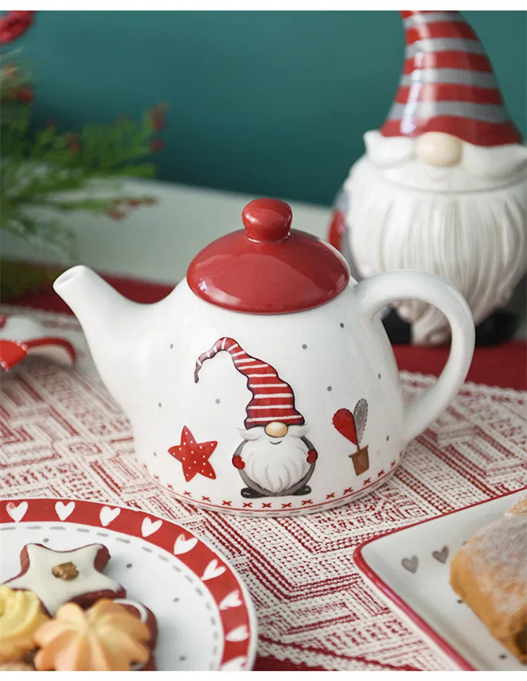A white ceramic teapot shaped like a Santa face is displayed on a red patterned cloth with food items.