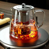 A clear borosilicate glass teapot with a stainless steel infuser filled with amber tea on a stovetop.