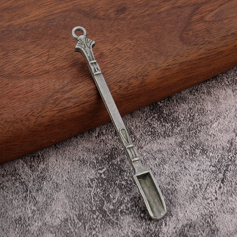 A silver metal mini shovel spoon with a loop handle rests on a gray textured surface.