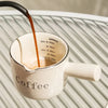 A stream of dark coffee is poured into a white ceramic cup on a striped surface.