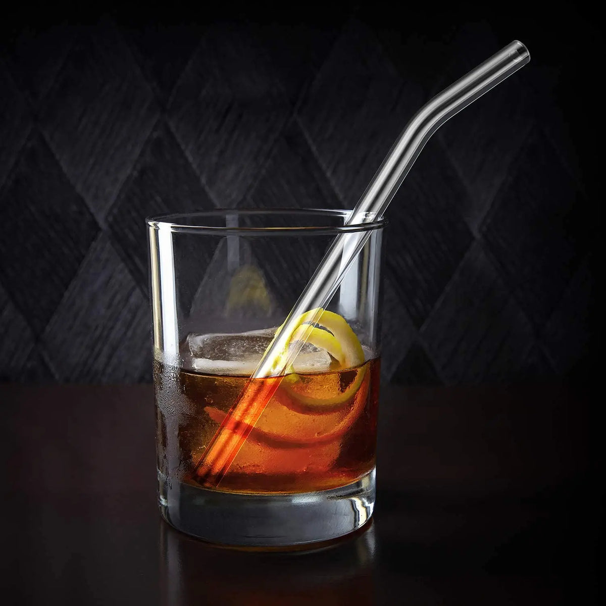 A clear glass contains an amber cocktail with a lemon garnish and is served with a clear drinking straw.