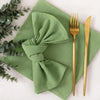 A set of reusable cotton cloth napkins in a sage green color with gold cutlery placed on a light surface.