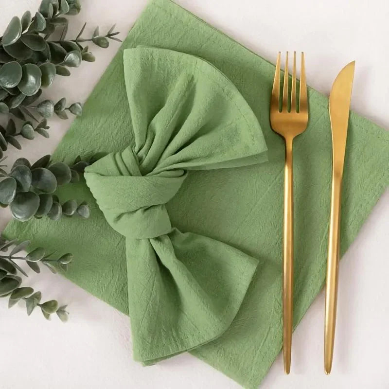 A set of reusable cotton cloth napkins in a sage green color with gold cutlery placed on a light surface.