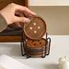 A hand is holding a round wooden coaster and a stack of coasters with an iron rack on a white surface.