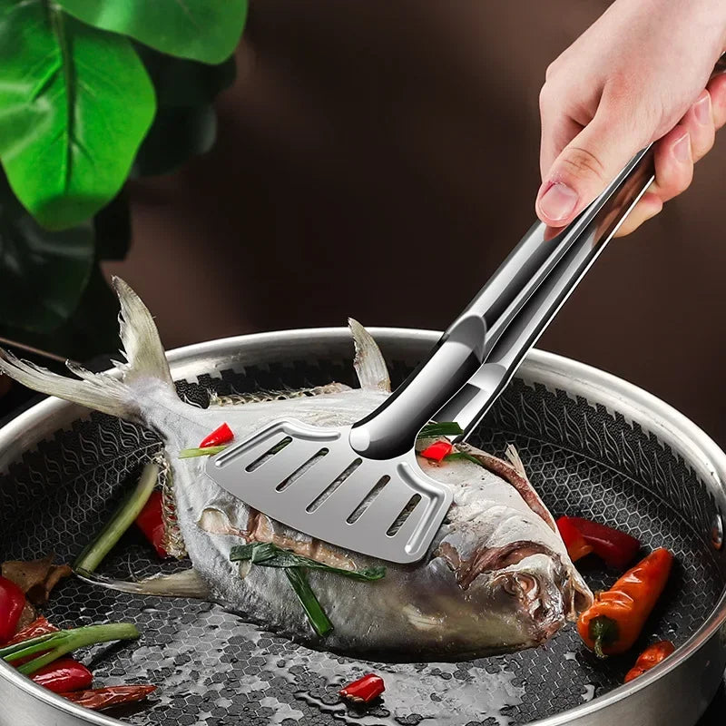 A hand uses stainless steel barbecue tongs to flip a whole fish in a frying pan.