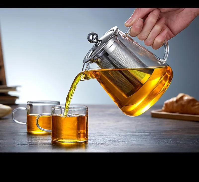 A hand pours amber tea from a glass teapot with a stainless steel infuser into a glass cup.