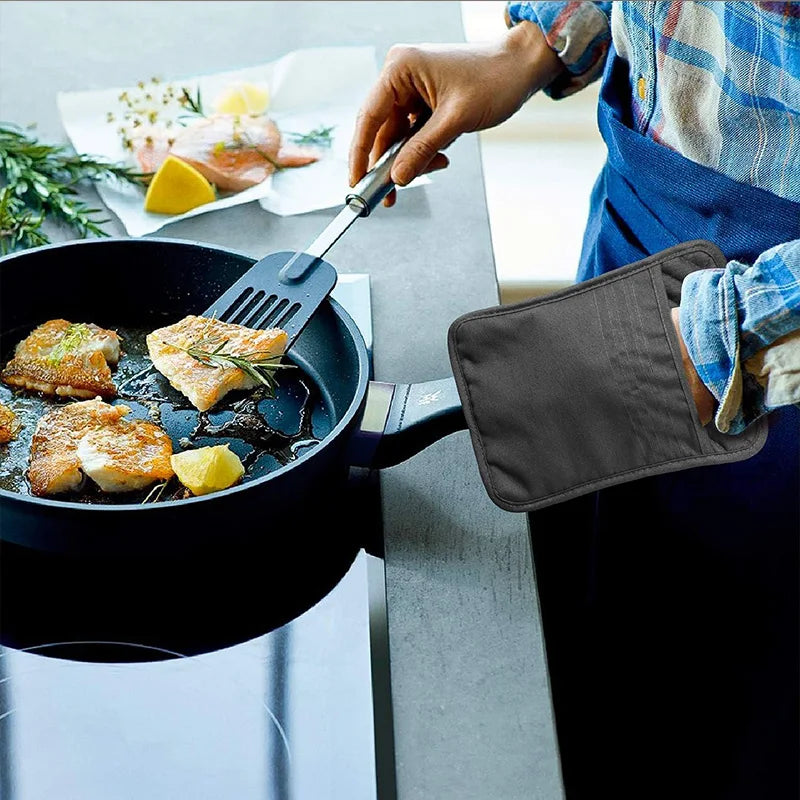A hand uses a spatula to lift cooked fish from a black pan on a stovetop, with a gray silicone cooking pad nearby.