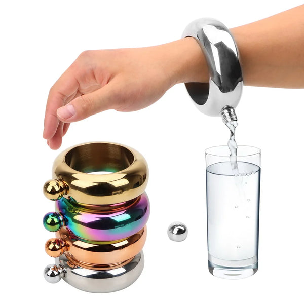 Eine Hand gießt Wasser aus einem silbernen Armband in ein Glas, neben einer Stapel bunter Metallringe.