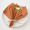 A set of ten rust-colored cotton table napkins is neatly folded and tied on a white plate with gold cutlery.