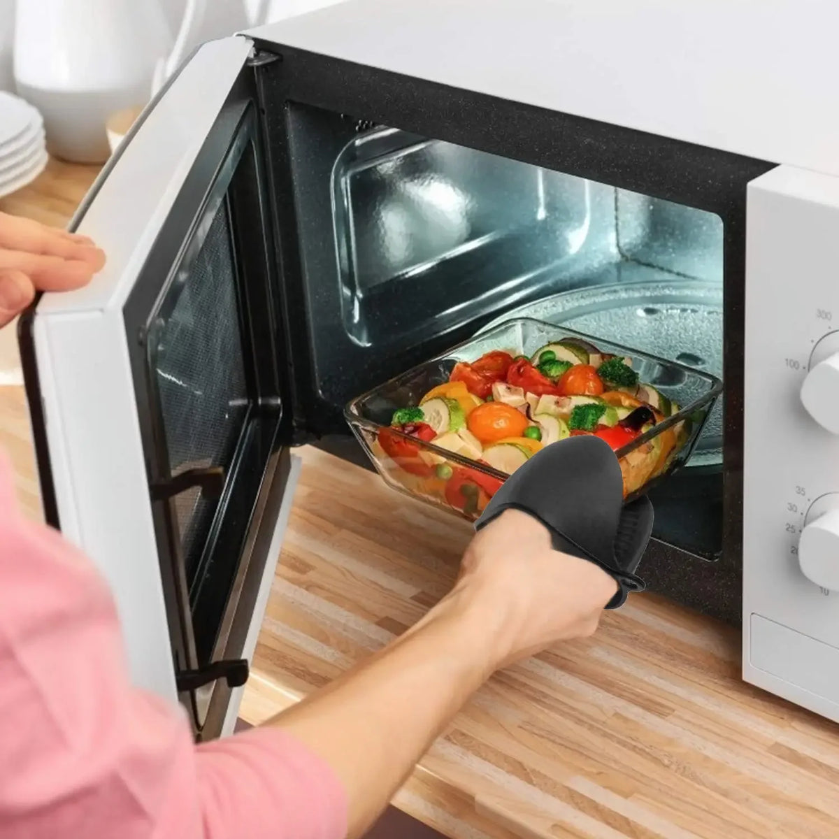A person is using a black silicone oven mitt to open and look inside a white electric oven.