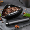 A black ceramic coffee dosing and spray kit with whole coffee beans in the bowl and a black applicator lying on a dark surfac