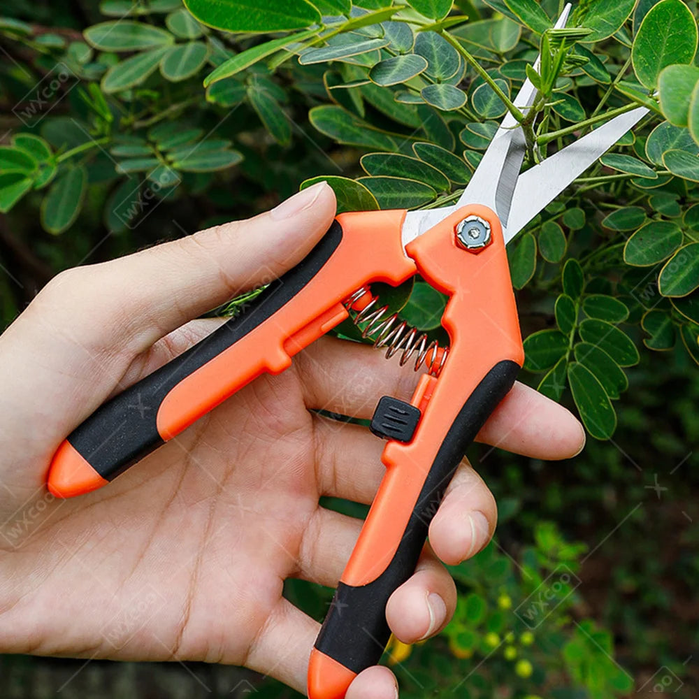 A hand holds a pair of orange and black professional stainless steel gardening shears against a green leafy background.