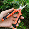 A hand holds a pair of orange and black professional stainless steel gardening shears against a green leafy background.