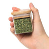 A hand holds an airtight glass spice jar with a bamboo lid filled with dried green herbs.