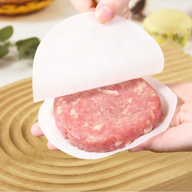 A hand holds a raw meat patty on a white baking paper sheet above a wooden cutting board.