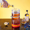A hand pours amber liquid from a glass bottle into a large glass dispenser filled with beer or wine.