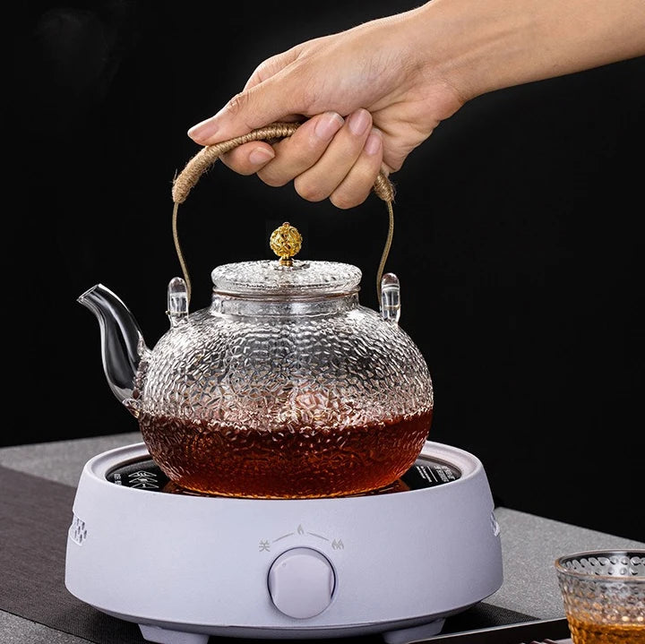 A hand is holding a hand-hammered glass teapot with a hemp rope design sitting on a white electric burner.