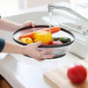 Hands are using a silicone splatter guard to pour liquid into a pot under a kitchen faucet.