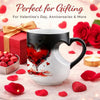 A white ceramic mug with a red heart design is displayed on a pink surface surrounded by red rose petals and a red gift box.