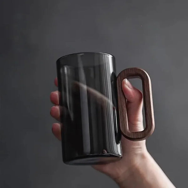 A hand is holding a dark smoke gray glass drinking cup with a natural wood handle against a gray background.